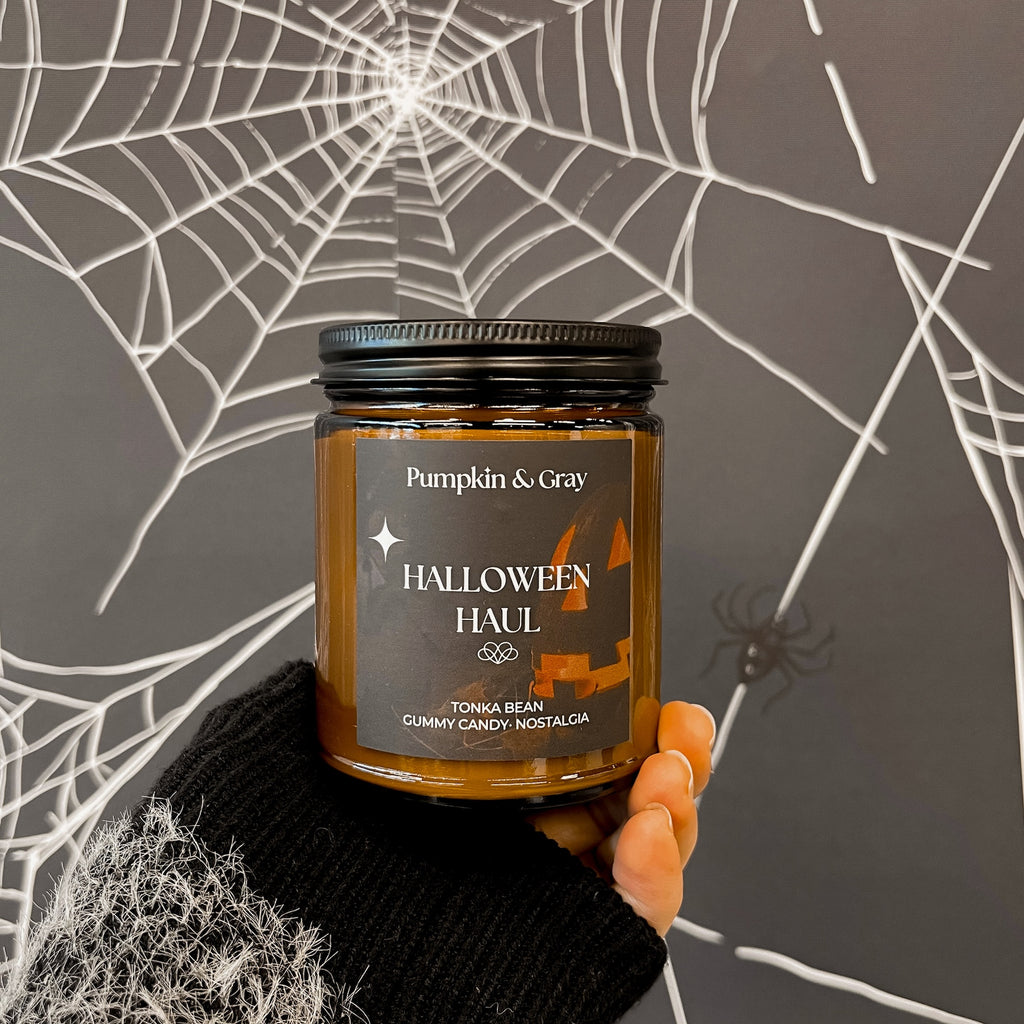 Candle labeled 'Pumpkin & Gray Halloween Haul' held against a spider web background