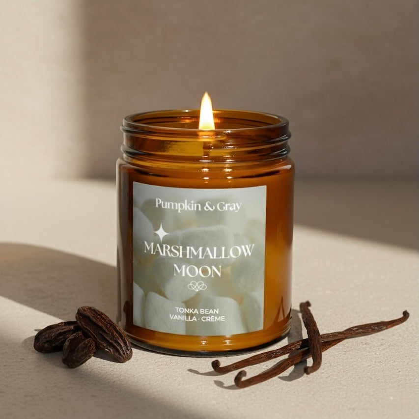 Candle in an amber jar labeled 'Marshmallow Moon' with vanilla beans and a vanilla pod on a neutral background.