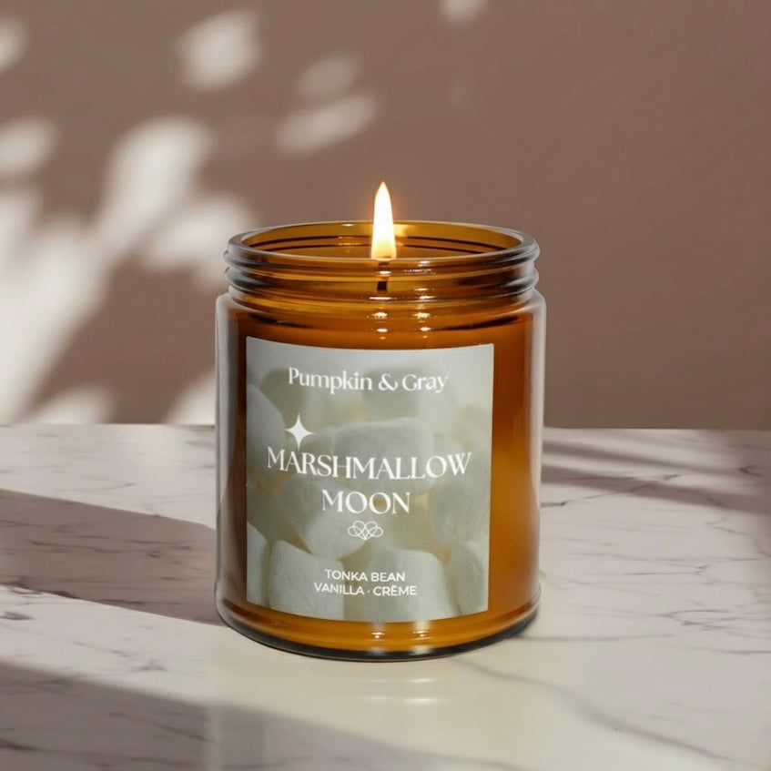 Candle in an amber jar labeled 'Marshmallow Moon' on a marble surface with a neutral background